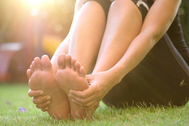 Πόνο στα πόδια Πόδι του ανθρώπου που κάθεται στο γρασίδι στο πάρκο κρατώντας τα πόδια του και τεντώστε τους μυς στο φως του ήλιου το πρωί.. - Φωτογραφία, εικόνα