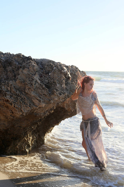 Ritratto a figura intera di donna dai capelli rossi che indossa abiti naufragati strappati. In piedi posa con le mani gestuali a roccioso oceano spiaggia paesaggio sfondo. - Foto, immagini