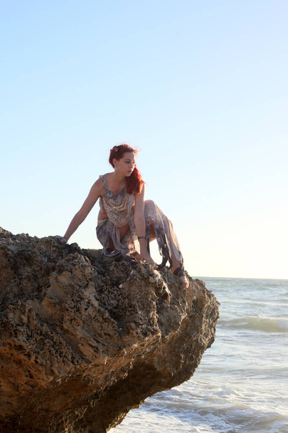 Teljes hosszúságú portré vörös hajú nőről, szakadt hajóroncsban. Álló póz geszturális kezek sziklás óceán strand táj háttér. - Fotó, kép