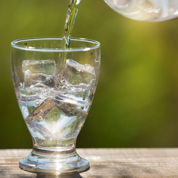 un bicchiere con cubetti di ghiaccio versando acqua da una bottiglia, all'aperto, luce solare e bokeh, il concetto di sete - Foto, immagini