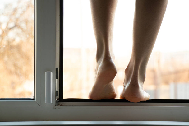 Ένα νεαρό κορίτσι στέκεται στο παράθυρο στο διαμέρισμά της, αυτοκτονώντας, πηδώντας τα παράθυρά τους, κατάθλιψη και αυτοκτονία ανθρώπων, με τα πόδια στο παράθυρο του σπιτιού. - Φωτογραφία, εικόνα