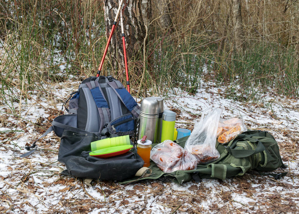 picture with snowy ground background and picnic accessories, backpacks, mugs, thermos and various dishes on the ground, tourist lunch at nature in winter - Foto, imagen