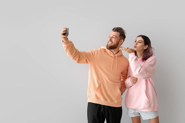 Cool pareja joven en sudaderas con capucha tomando selfie sobre fondo claro - Foto, Imagen