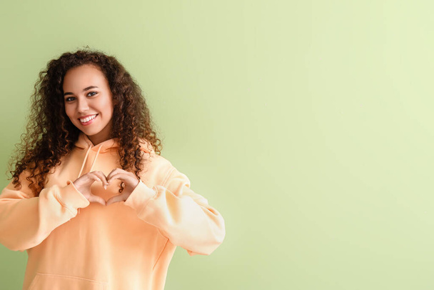 Beautiful young African-American woman in stylish hoodie making heart with her hands on color background - Photo, Image