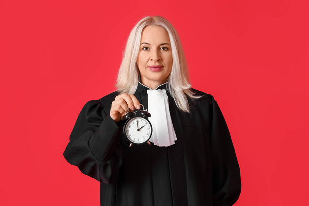 Ώριμη γυναίκα δικαστής με ρόμπα με ξυπνητήρι σε κόκκινο φόντο - Φωτογραφία, εικόνα