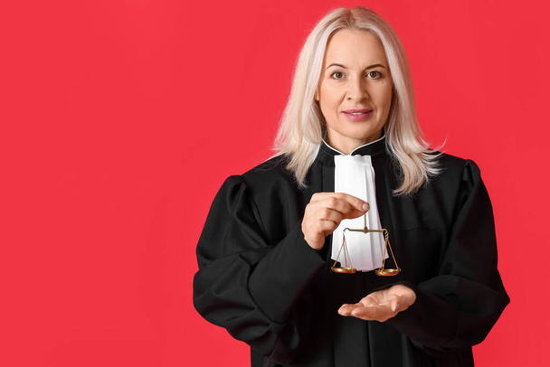 Ώριμη γυναίκα δικαστής με ρόμπα με ζυγαριές δικαιοσύνης σε κόκκινο φόντο - Φωτογραφία, εικόνα