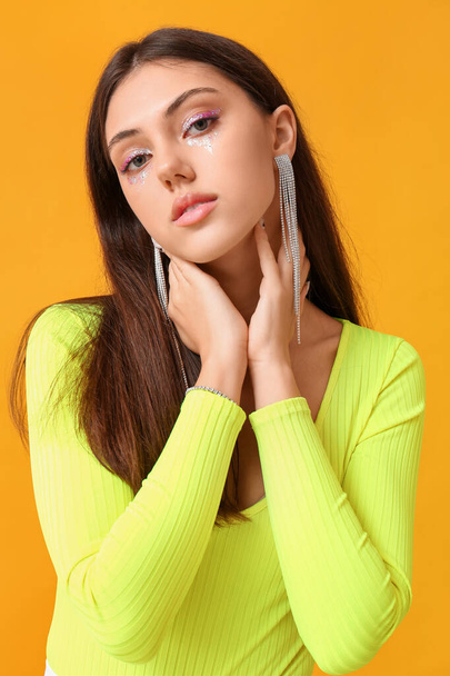 Woman with creative makeup and stylish jewelry touching neck on yellow background - Foto, afbeelding