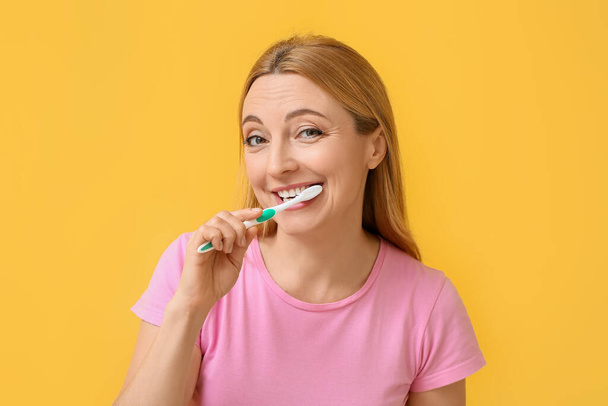 Beautiful mature woman brushing teeth on yellow background - Fotoğraf, Görsel