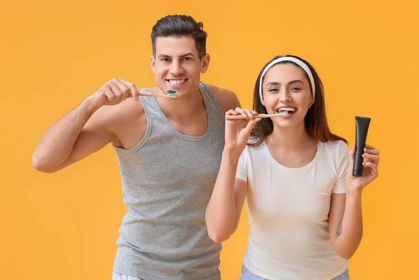 Νεαρό ζευγάρι βουρτσίζει τα δόντια με ενεργό πάστα δοντιών άνθρακα σε κίτρινο φόντο - Φωτογραφία, εικόνα