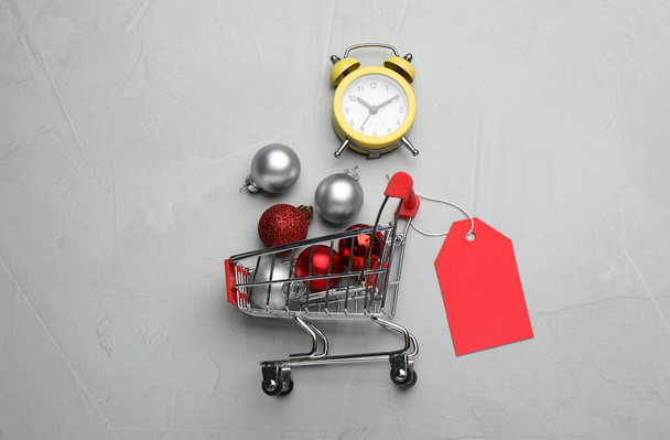 Shopping cart with Christmas decor and alarm clock on grey background, flat lay. Sale concept - Foto, Imagem