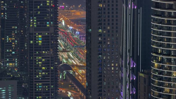 Highway and overpass between skyscrapers with glowing windows in Dubai downtown aerial night timelapse. Glowing windows in towers near metro station and busy traffic from above. - Φωτογραφία, εικόνα