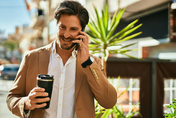 若いヒスパニック系のビジネスマンが市内のスマートフォンで話してコーヒーを飲む. - 写真・画像