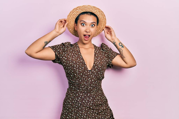 Hermosa mujer hispana con el pelo corto usando sombrero de verano asustada y conmocionada con la sorpresa y la expresión asombrada, miedo y cara emocionada.  - Foto, Imagen