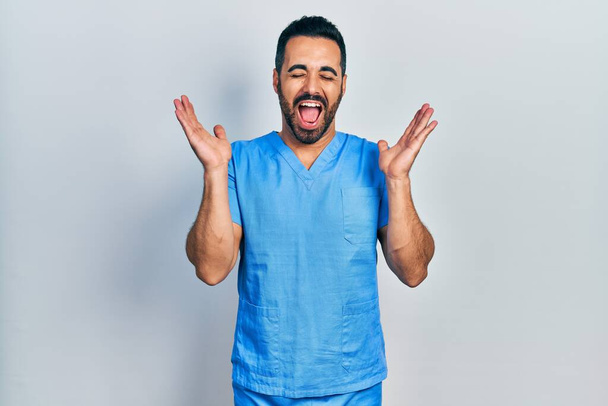 Knappe Spaanse man met baard in blauw verpleegstersuniform die gek en gek viert op succes met opgeheven armen en gesloten ogen schreeuwend opgewonden. winnaarsconcept  - Foto, afbeelding