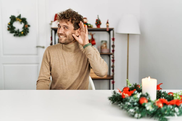 Fiatal, jóképű, szakállas férfi ül az asztalon karácsonyi dekorációval, mosolygva, fültől fülig hallgatva egy pletykát vagy pletykát. süketség fogalma.  - Fotó, kép