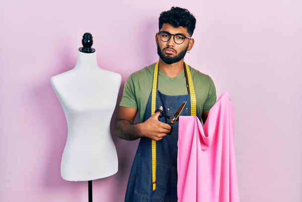 Arab man with beard dressmaker designer holding scissors and cloth clueless and confused expression. doubt concept.  - Photo, image