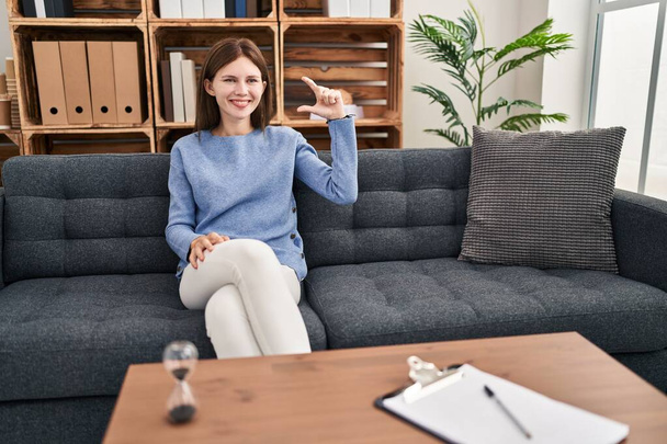 Young brunette woman at consultation office smiling and confident gesturing with hand doing small size sign with fingers looking and the camera. measure concept.  - 写真・画像