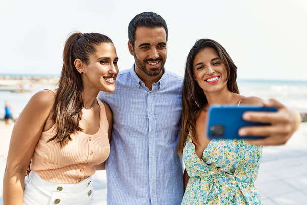 Tres jóvenes amigos hispanos sonriendo felices hacen selfie junto al smartphone en la playa. - Foto, Imagen