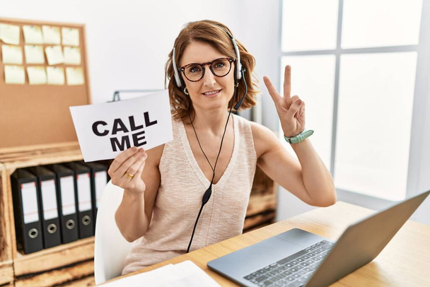 Middle age brunette woman wearing operator headset holding call me banner smiling looking to the camera showing fingers doing victory sign. number two.  - Фото, изображение