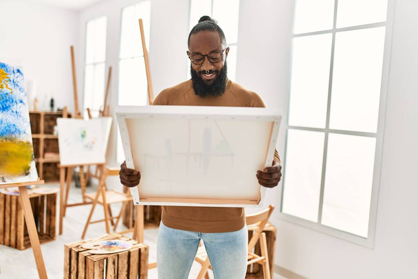 Jovem artista afro-americano homem sorrindo feliz segurando tela no estúdio de arte. - Foto, Imagem