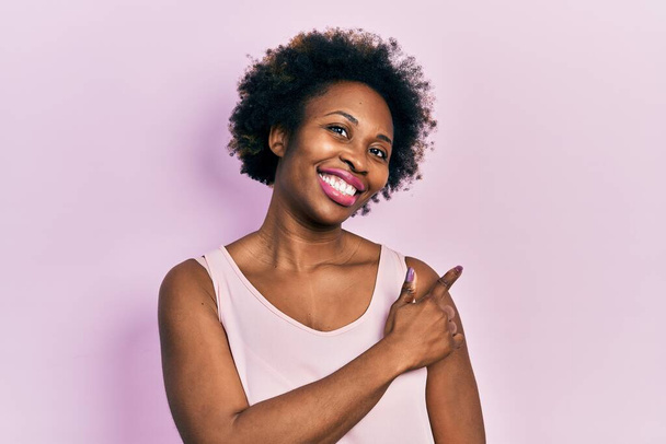Молода афроамериканська жінка у звичайній сорочці без рукавів весела з посмішкою обличчя, що вказує рукою і пальцем на бік з радісним і природним виразом обличчя  - Фото, зображення