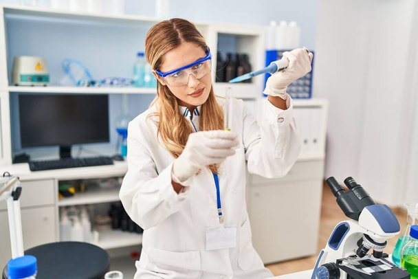 Mujer rubia joven vistiendo uniforme científico vertiendo líquido usando pipeta en el laboratorio - Foto, Imagen