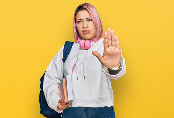 Femme hispanique aux cheveux roses portant un sac à dos d'étudiant et des écouteurs arrêtant de chanter avec la paume de la main. expression d'avertissement avec geste négatif et sérieux sur le visage.  - Photo, image