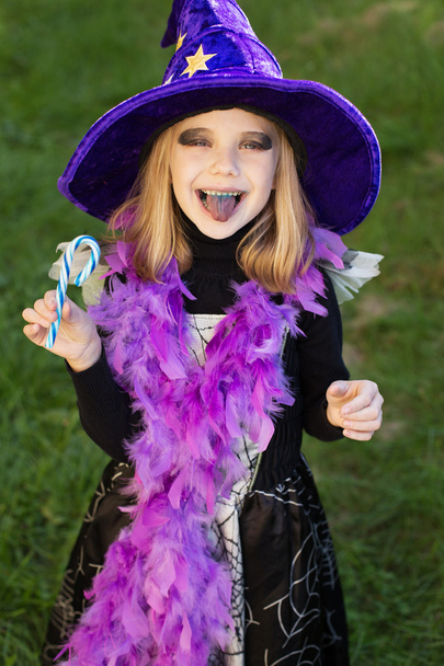 Маленька красива дівчинка з костюмом Хеллоуїн відьми їсть цукерки і показує фіолетовий язик
 - Фото, зображення