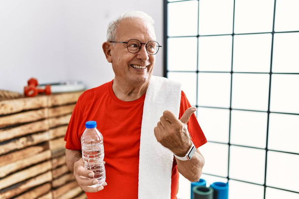 Homme âgé portant des vêtements de sport et serviette à la salle de gym pointant vers le dos derrière avec la main et les pouces levés, souriant confiant  - Photo, image