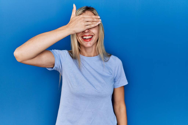 Krásná blondýnka na sobě ležérní tričko přes modré pozadí s úsměvem a smíchem s rukou na tváři zakrývající oči pro překvapení. slepý koncept.  - Fotografie, Obrázek
