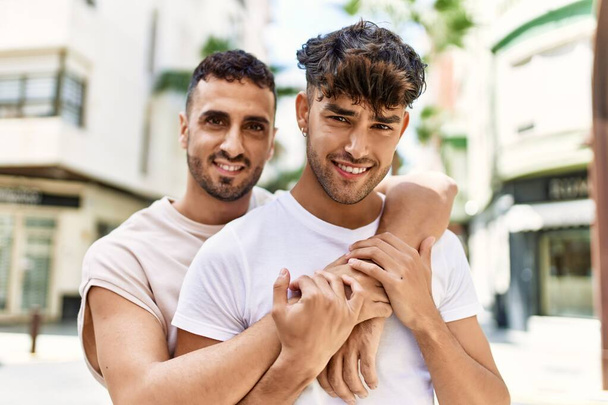 Два латиноамериканских мужчины улыбаются уверенно обнимая друг друга стоя дома - Фото, изображение