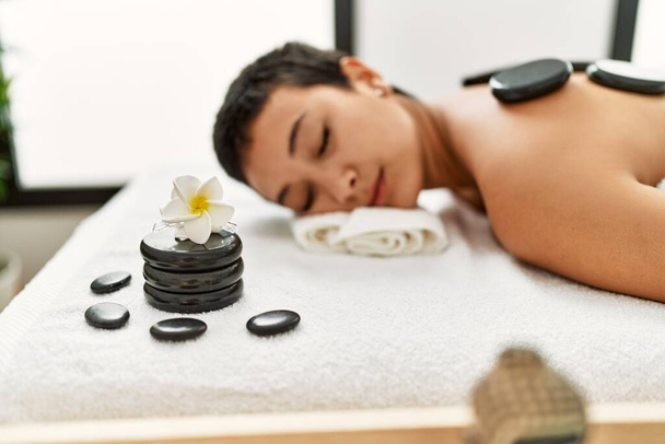 Mujer hispana joven teniendo masaje de espalda usando piedras calientes negras en el centro de belleza - Foto, Imagen