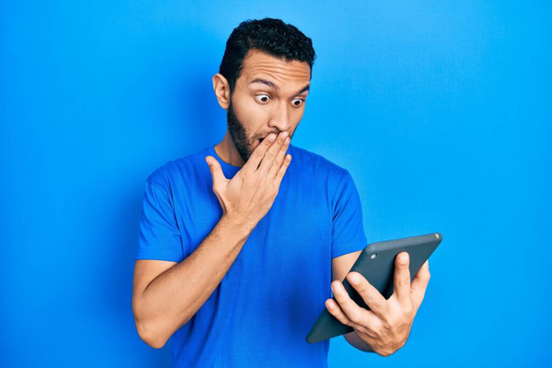 Hispanischer Mann mit Bart mit Touchpad-Gerät, das Mund und Hand bedeckt, schockiert und ängstlich vor Fehlern. Überraschte Miene  - Foto, Bild