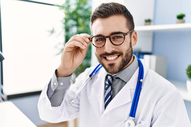 Giovane uomo ispanico indossa uniforme medico e occhiali in clinica - Foto, immagini