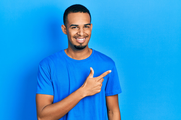 青いシャツを着た若いアフリカ系アメリカ人の男性は、笑顔で笑顔を見せ、手と指を顔に喜びと自然な表情で横に向けます。  - 写真・画像