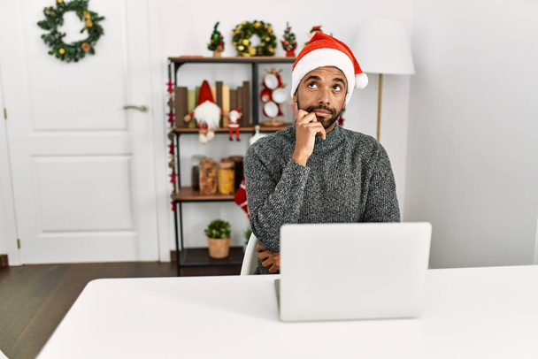 Jeune homme hispanique avec barbe portant un chapeau de Noël en utilisant un ordinateur portable visage sérieux penser à la question avec la main sur le menton, réfléchi sur l'idée déroutante  - Photo, image