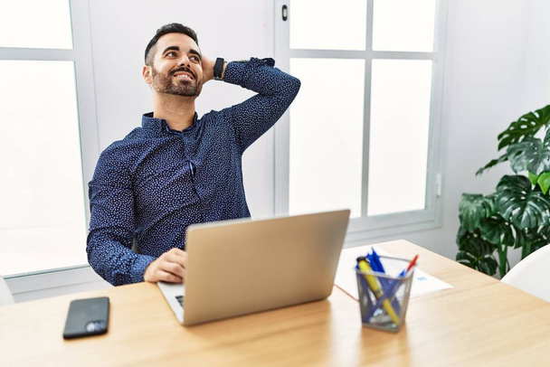 Junge hispanische Mann mit Bart arbeitet im Büro mit Laptop lächelnd zuversichtlich berühren Haare mit Hand nach oben Geste, posiert attraktiv und modisch  - Foto, Bild