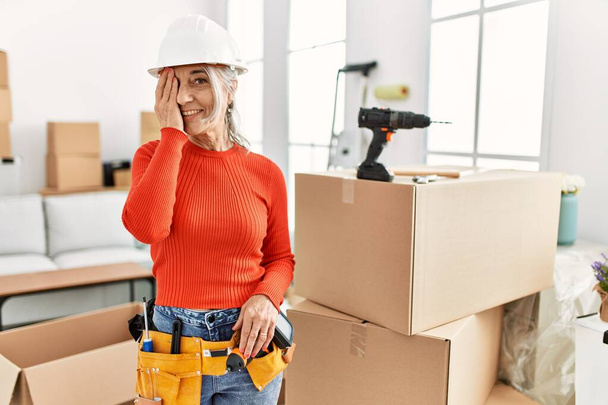 Middle age grey-haired woman wearing hardhat standing at new home covering one eye with hand, confident smile on face and surprise emotion.  - Photo, Image