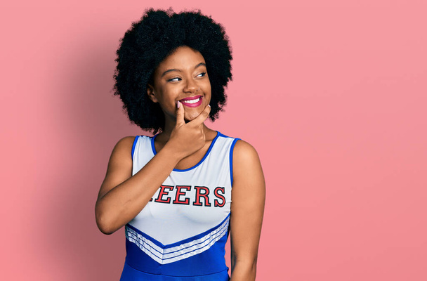 Jonge Afro-Amerikaanse vrouw in cheerleader uniform met de hand op de kin denkend aan vragen, dure uitdrukking. glimlachend met een bedachtzaam gezicht. twijfelconcept.  - Foto, afbeelding