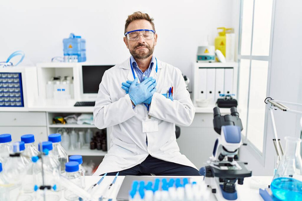 Middle age man working at scientist laboratory smiling with hands on chest with closed eyes and grateful gesture on face. health concept.  - Photo, Image