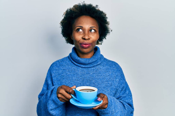 Fiatal afro-amerikai nő iszik egy csésze kávét mosolyog oldalra néz, és félrenéz gondolkodás közben..  - Fotó, kép