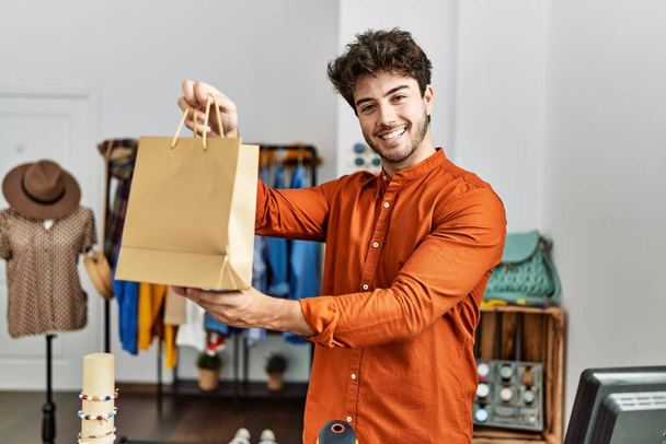 młody Latynoski sklepikarz człowiek uśmiecha szczęśliwy trzymając torbę na zakupy w sklepie odzieżowym. - Zdjęcie, obraz