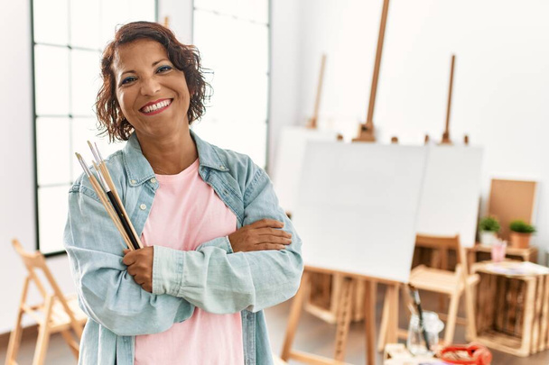 Mujer artista hispana de mediana edad sonriendo feliz de pie con los brazos cruzados gesto en el estudio de arte. - Foto, imagen