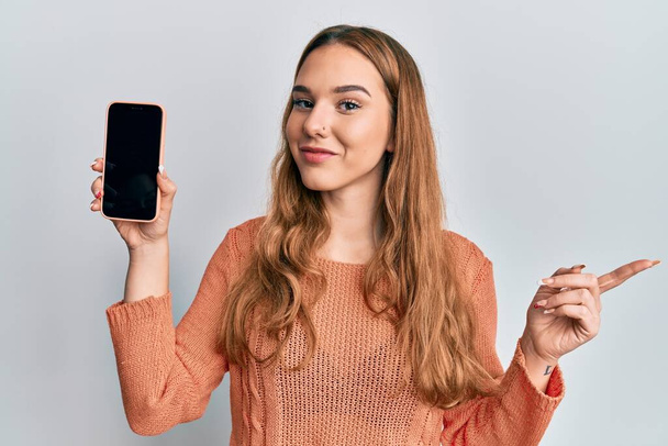 Young blonde woman holding smartphone showing screen smiling happy pointing with hand and finger to the side  - Фото, зображення