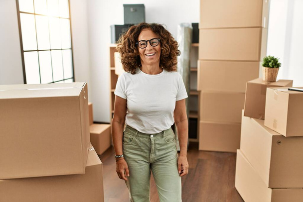 Mujer hispana de mediana edad vistiendo ropa casual de pie alrededor de cajas de cartón en un nuevo hogar luciendo positiva y feliz de pie y sonriendo con una sonrisa confiada mostrando los dientes  - Foto, Imagen