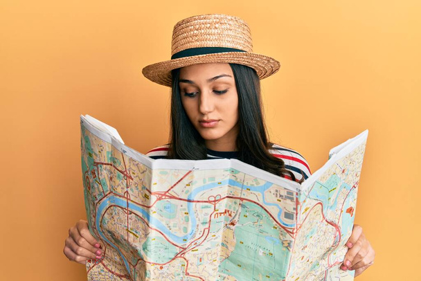 Young hispanic woman wearing summer hat holding map relaxed with serious expression on face. simple and natural looking at the camera.  - Foto, Imagem