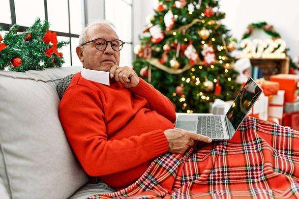 Hombre mayor con el pelo gris usando el ordenador portátil sentado junto a la cara seria árbol de Navidad pensando en la pregunta con la mano en la barbilla, pensativo acerca de la idea confusa  - Foto, Imagen