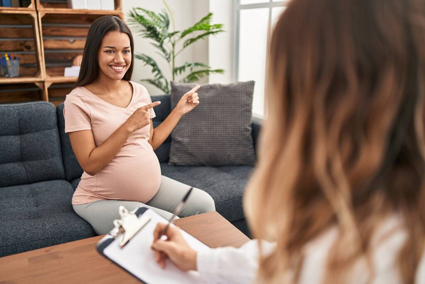 Młoda kobieta w ciąży na sesji terapeutycznej uśmiechnięta i patrząca w kamerę skierowaną obiema rękami i palcami do boku.  - Zdjęcie, obraz