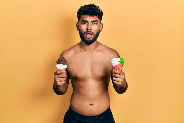 Homem árabe com barba vestindo roupa de banho comendo dois cones de sorvete com medo e chocado com a expressão surpresa e surpreso, medo e rosto animado.  - Foto, Imagem