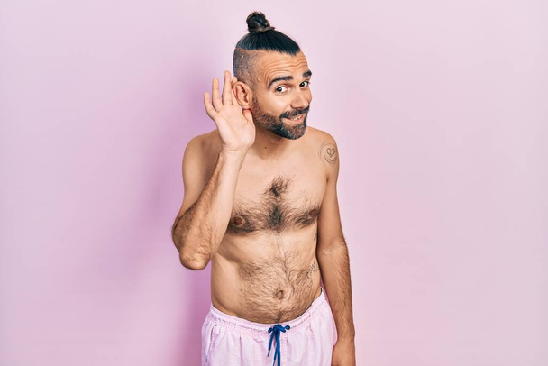 Jonge Spaanse man zonder shirt in een zwempak glimlachend met hand op oor luisterend naar geruchten of roddels. doofheid.  - Foto, afbeelding